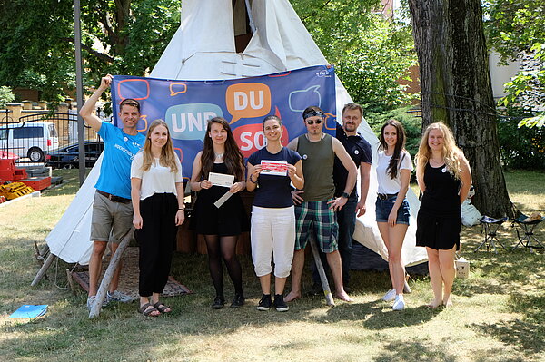 Auf dem Bild steht eine Gruppe von Personen vor einem Tipi und hält ein Banner mit der Aufschrift UND DU SO? hoch. In der Mitte halten zwei Studentinnen Gutscheine zur Kamera.  