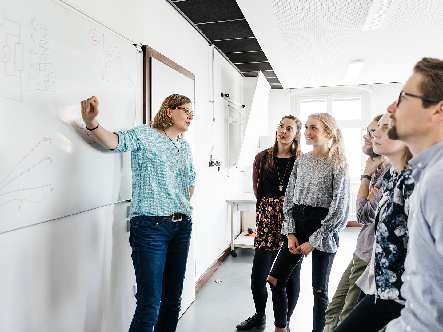 Eine Lehrerin erklärt an einer weißen Tafel physikalische Diagramme und Formeln, umgeben von sechs jungen Erwachsenen, die aufmerksam zuhören. Die Lehrerin trägt eine Brille, eine türkisfarbene Bluse und Jeans. Die Tafel zeigt handgezeichnete Diagramme, darunter Kurven und ein Schaltbild. Im Hintergrund befindet sich ein heller Raum mit hohen Fenstern und moderner Ausstattung.
