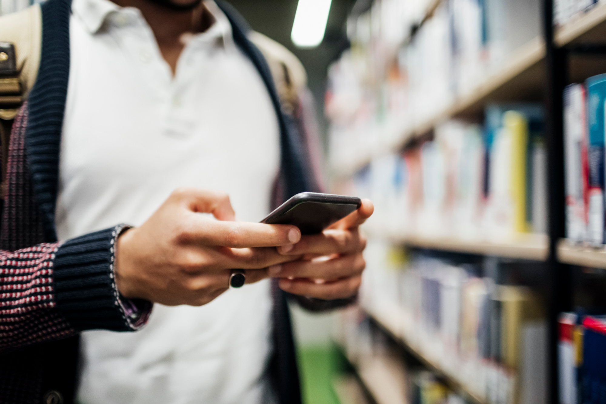 Studierender steht neben einem Bücherregal und hält ein Smartphone in der Hand.
