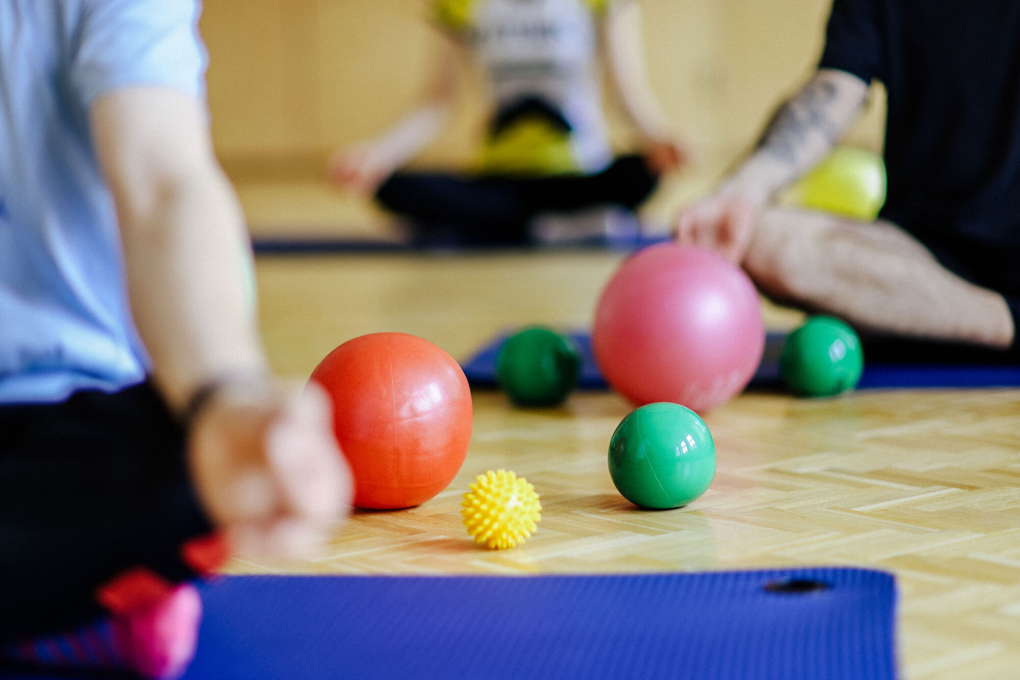 Verschiedene bunte Gymnastikbälle liegen auf Holzboden vor blauen Matten, umgeben von meditierenden Personen im Schneidersitz.